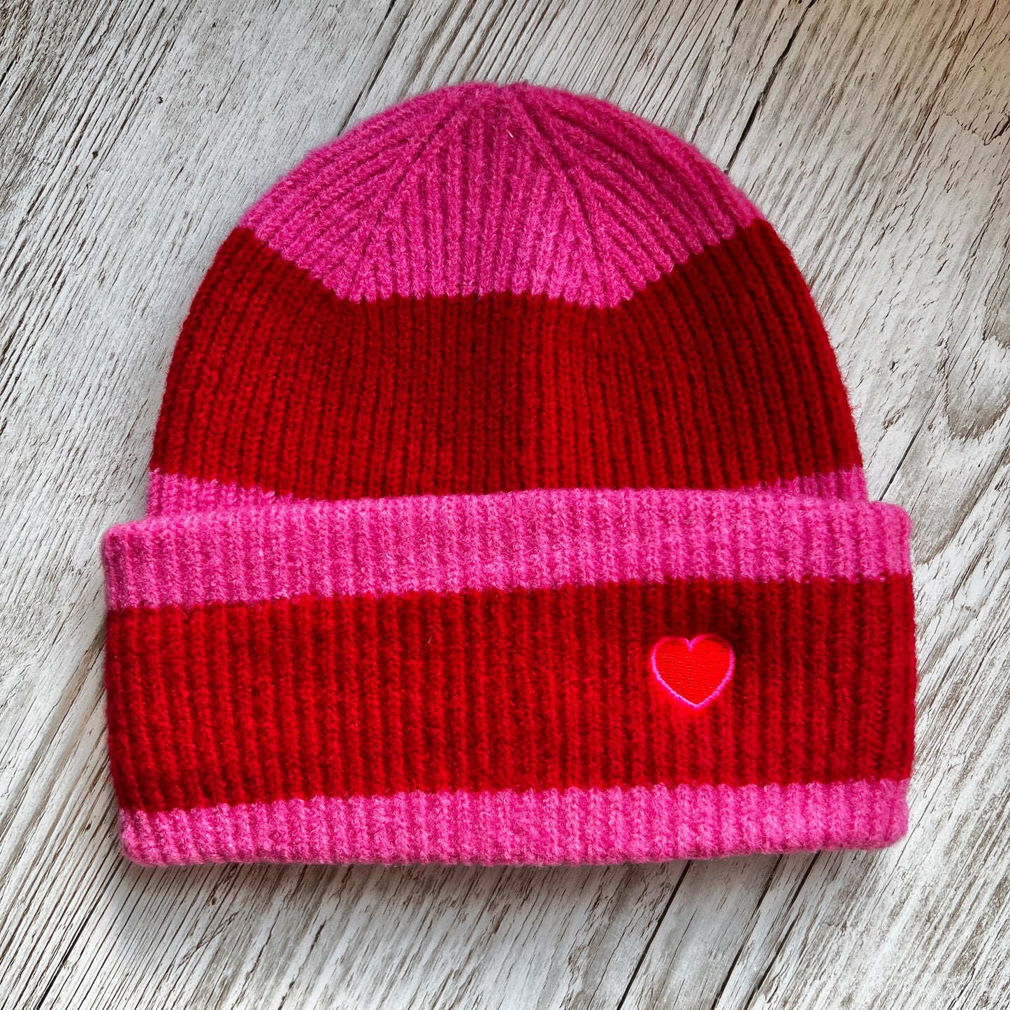 Red and pink striped beanie with an embroidered pink and red heart on the cuff, on a wooden background.