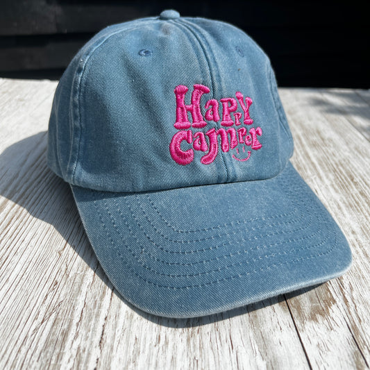 blue dad cap with pink embroidered 'Happy Camper' in fun lettering, on w light wooden background