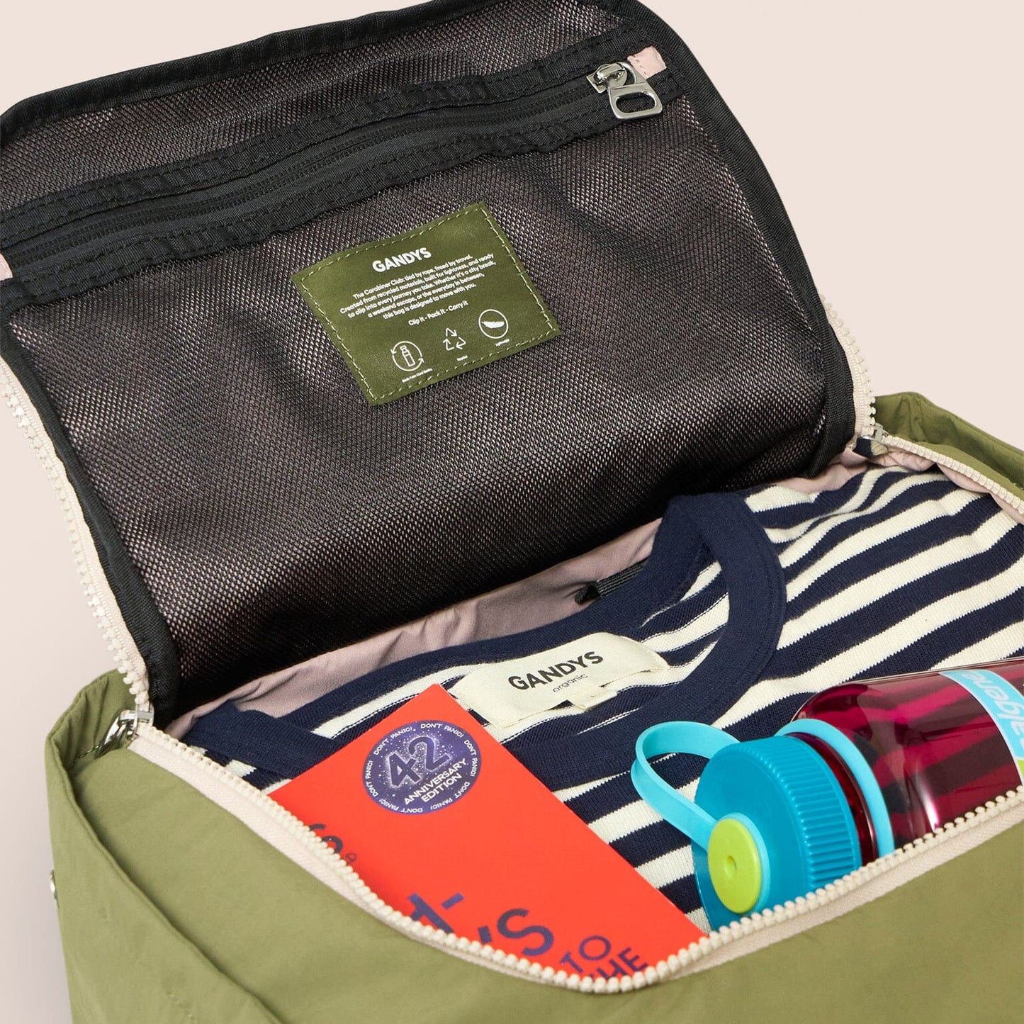 Green travel bag with GANDYS branding, containing a red book, striped towel, and colorful sippy cup.