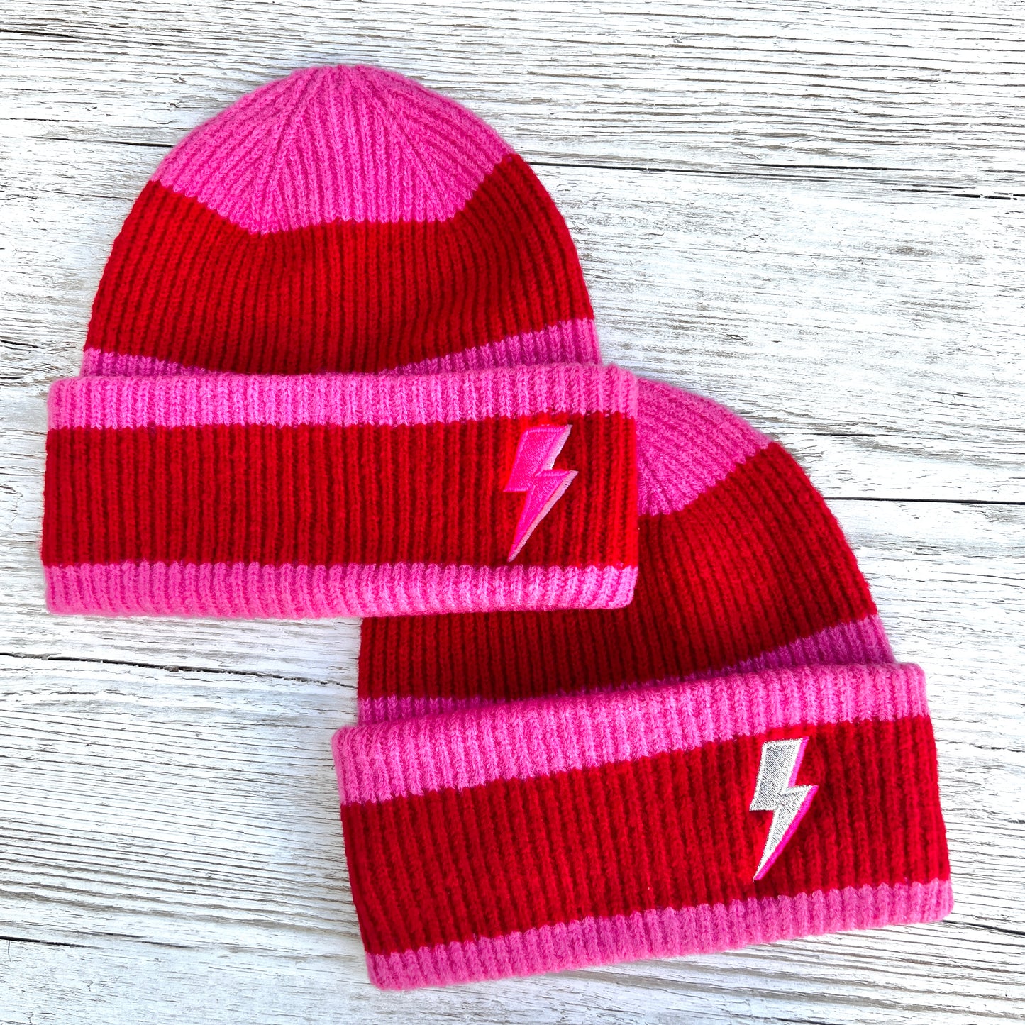 Two Red and pink stripped beanies with pink and silver embroidered bolts on a wooden background