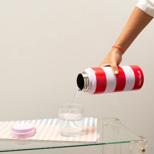 Person pouring water from a red and lilac striped thermos into a glass on a pastel striped matt at on a glass table