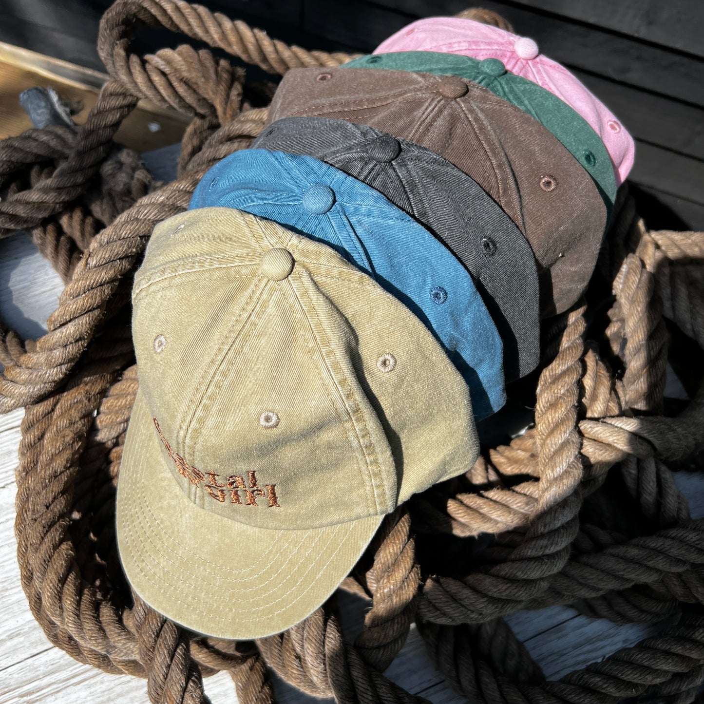 six coastal cowgirl embroidered caps of varying colours stacked together sitting on top of a pile of rope