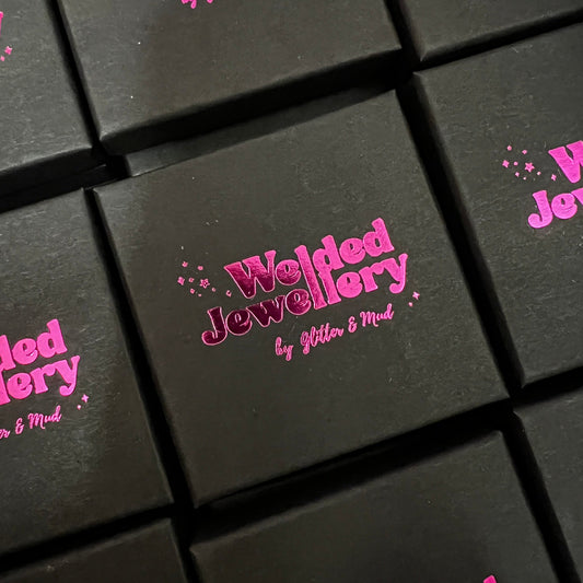 A boxed gift with a pink metallic and black design, featuring the text 'Welded Jewellery by Glitter & Mud'.