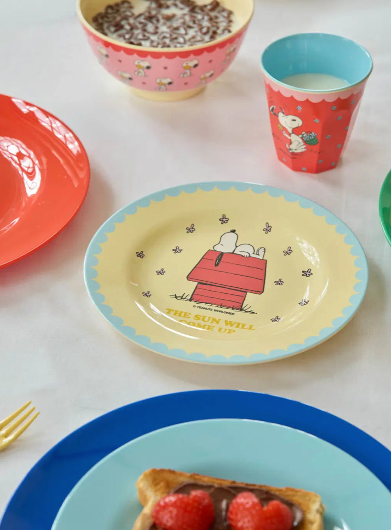 A colorful dinnerware set featuring a central yellow plate with Snoopy in a red doghouse, surrounded by a light blue scalloped rim. Other visible pieces include a red cup with a cartoon character, a pink bowl, and other plates on a white table.