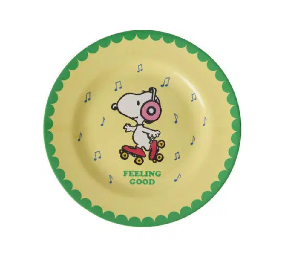 A round yellow plate with a green scalloped rim, featuring a cartoon dog character riding a pink scooter and the words 'FEELING GOOD' printed below, on a white background.