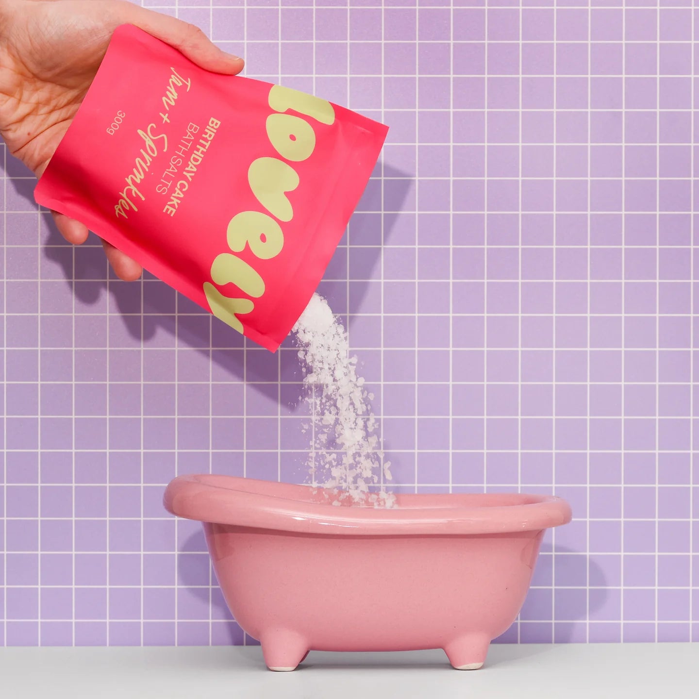 Person pouring a packet of bath salts into a small pink bathtub against a purple tiled wall.
