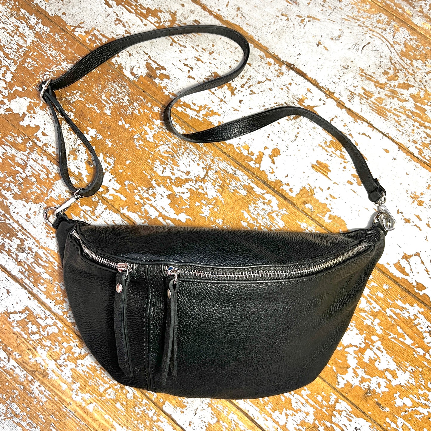 A black leather bumbag with a detachable strap, zippered compartments, and silver coloured fixings on a wooden background.