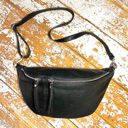 A black leather bumbag with a detachable strap, zippered compartments, and silver coloured fixings on a wooden background.