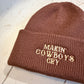 Brown beanie with embroidered  'MAKIN COWBOYS CRY' text on a wooden surface