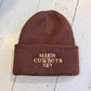Brown beanie with embroidered 'Makin' Cowboys Cry' text on a light wooden surface