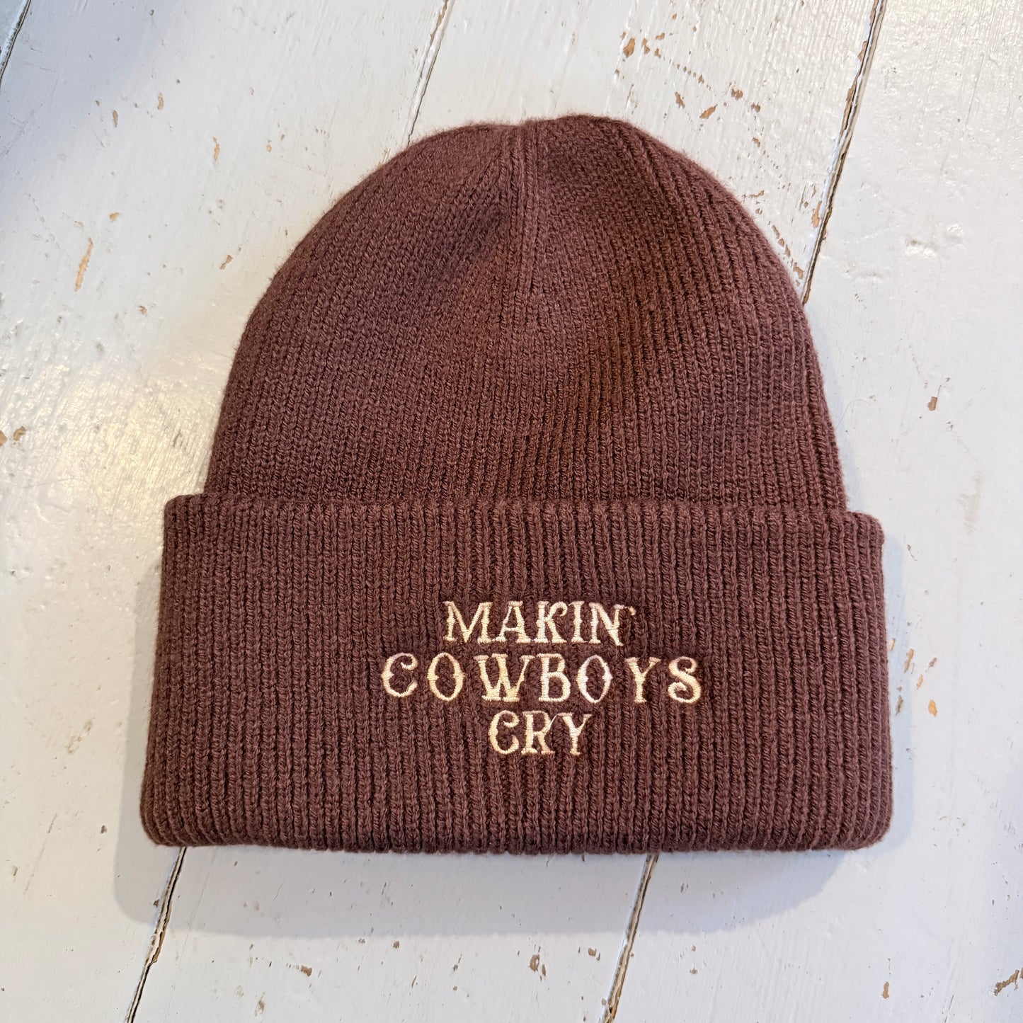 Brown beanie with embroidered 'Makin' Cowboys Cry' text on a light wooden surface
