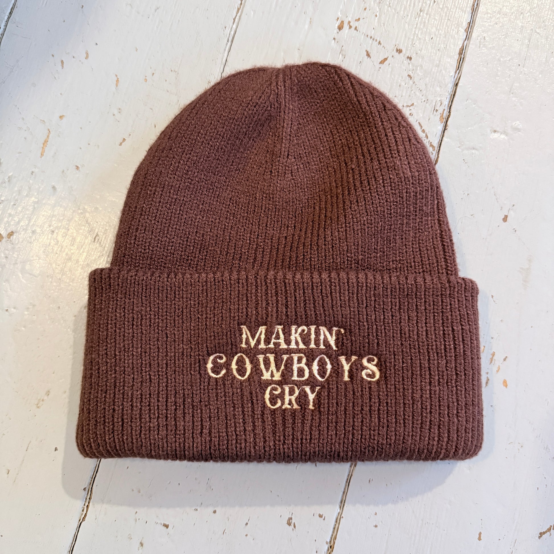 Brown beanie with embroidered 'Makin' Cowboys Cry' text on a light wooden surface