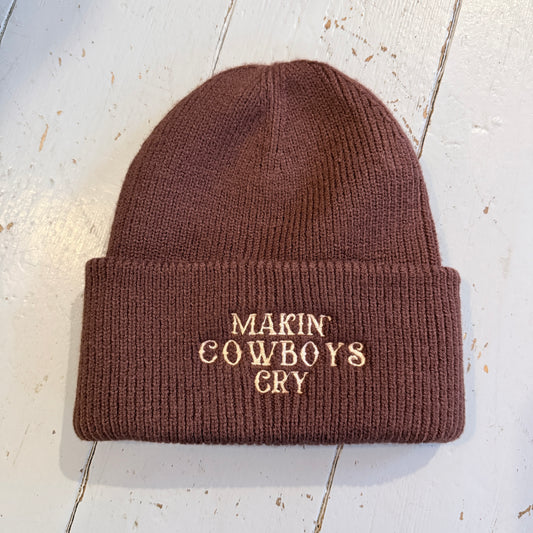 Brown beanie with embroidered 'Makin' Cowboys Cry' text on a light wooden surface