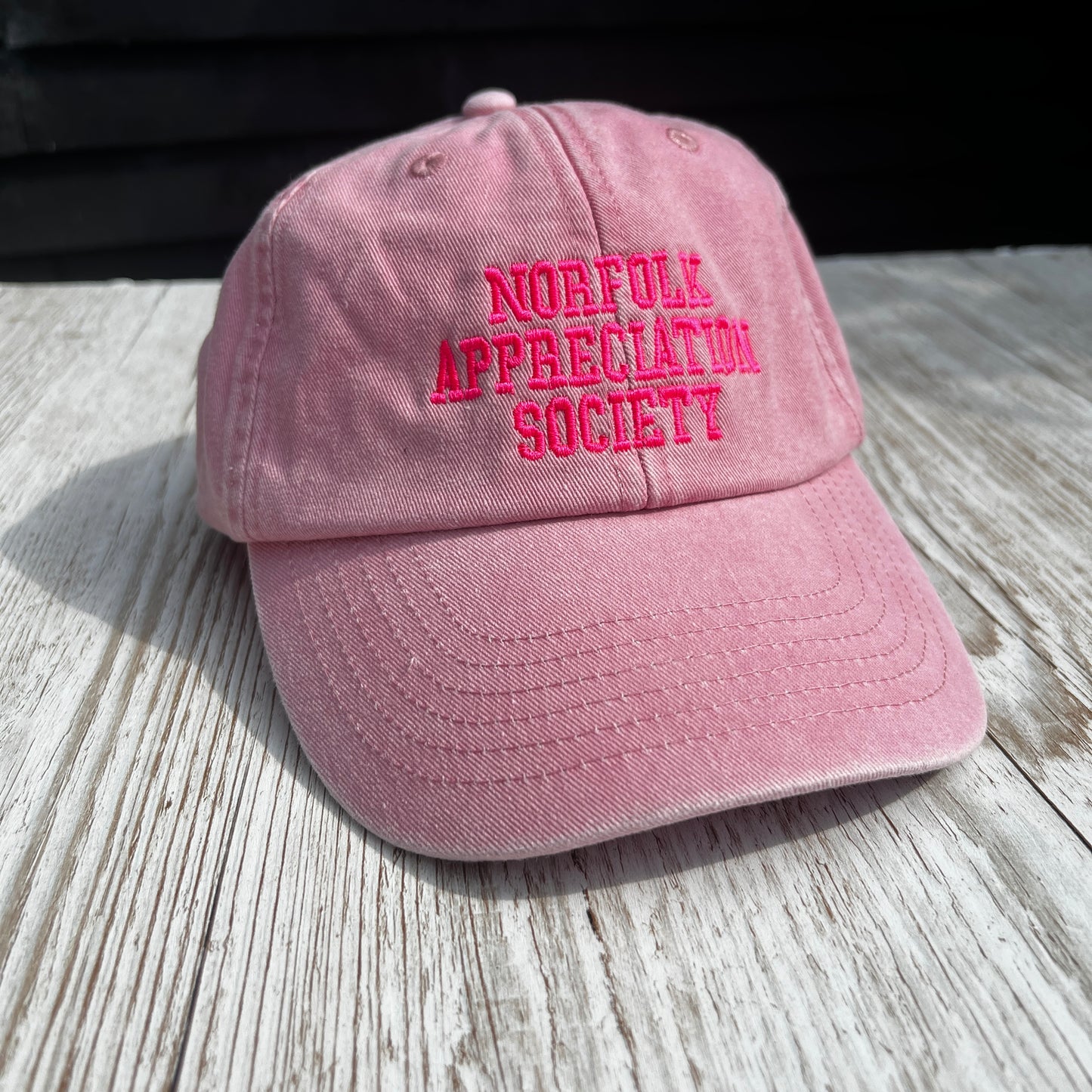 Embroidered washed pink dads cap 'Norfolk appreciation society' on a wooden surface