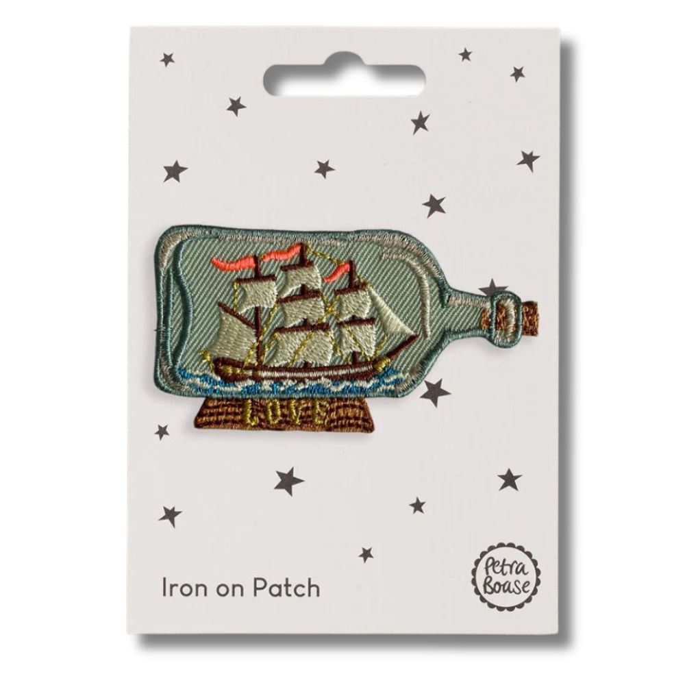 Embroidered  patch depicting a ship in a bottle with embroidered details in blue, gold, and multicolor, branded by Petra Boase, with an 'iron on' backing card.