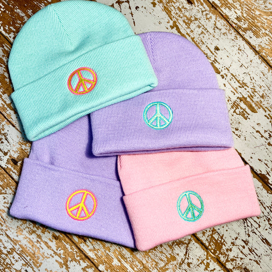 Four pastel-colored beanie hats with embroidered peace signs/ CND's on them, laid out on a wooden surface.