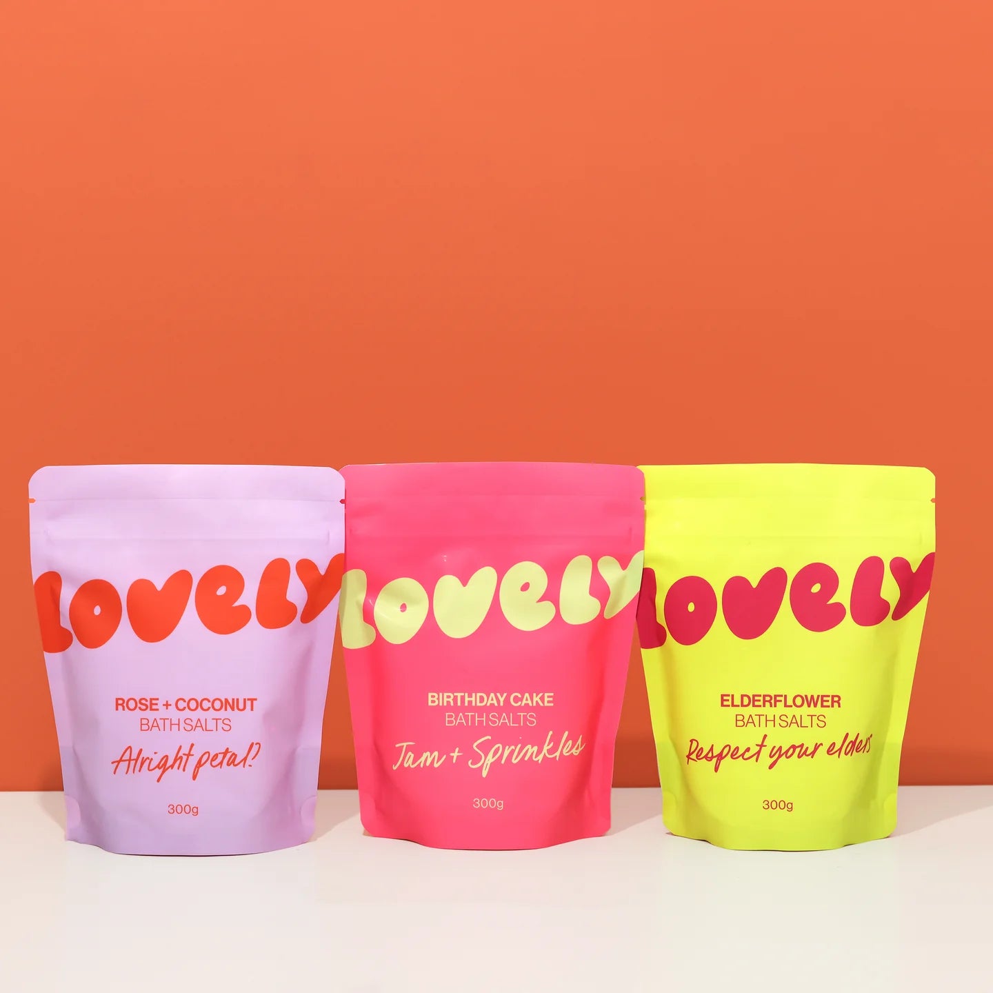 Three 'lovely' branded bath salt packages in different colors with text on an orange background.
