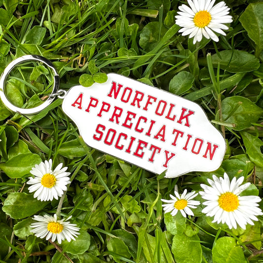An enamel red and white  keyring with 'NORFOLK APPRECIATION SOCIETY' text on it, placed on a grassy surface with daisies around.