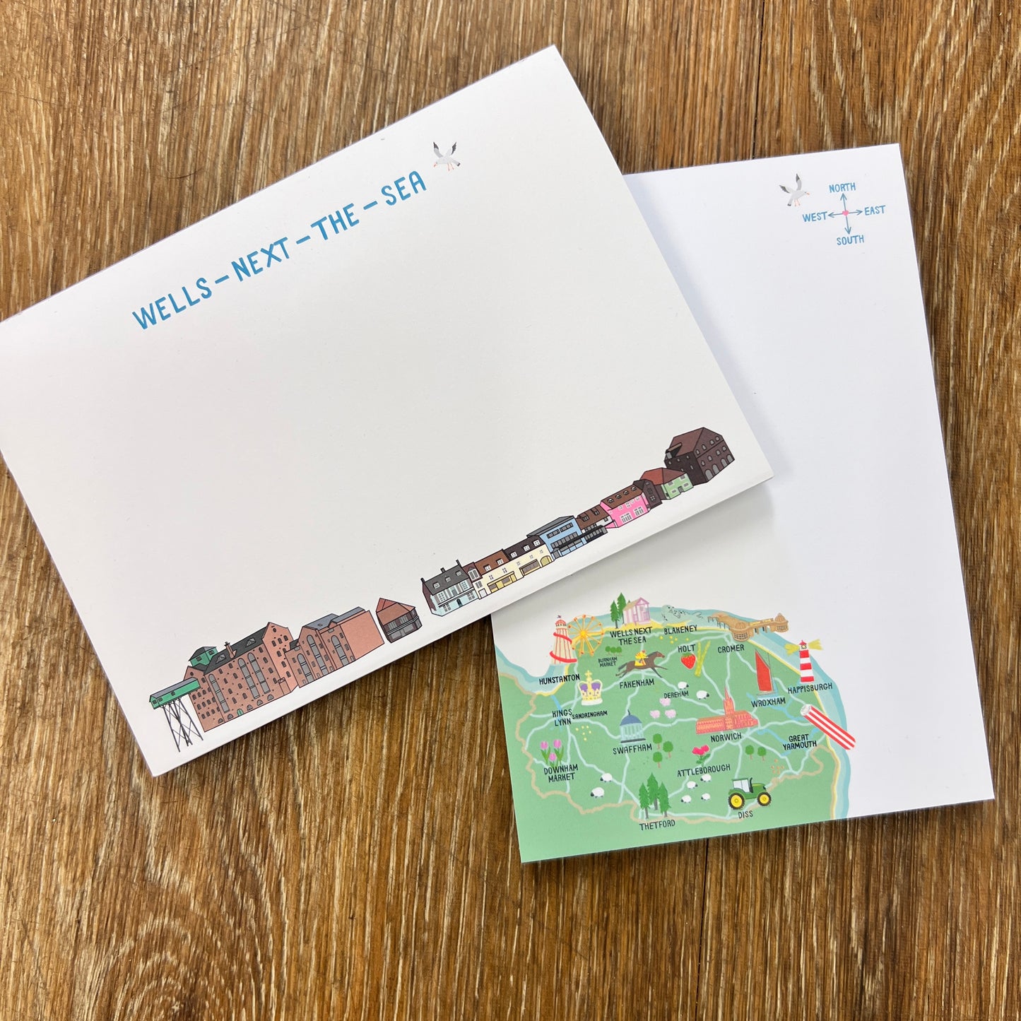 Two note pads on a wooden surface, one with wells next-the sea Quay illustrated buildings and the other with a map of the North Norfolk coast .