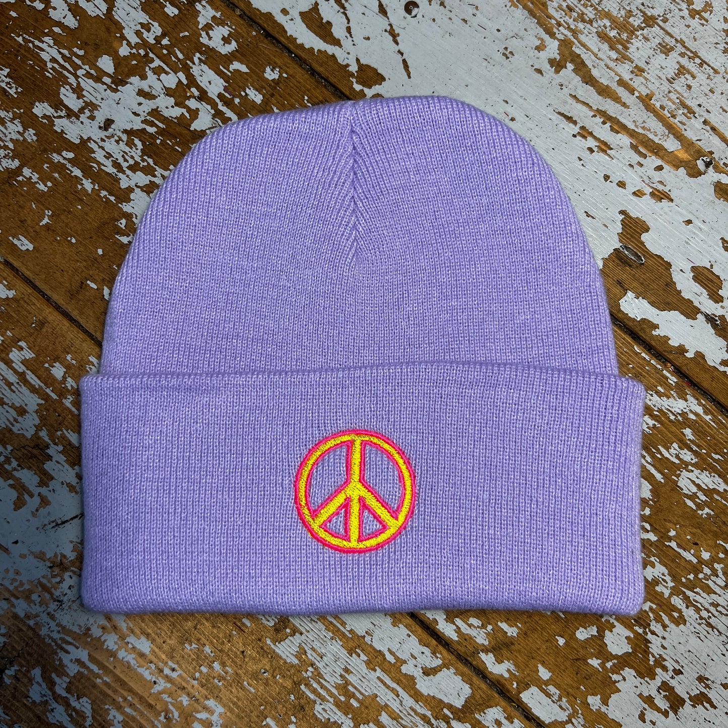 Pastel Lilac beanie hat with embroidered peace sign on cuff , laid out on a wooden surface.