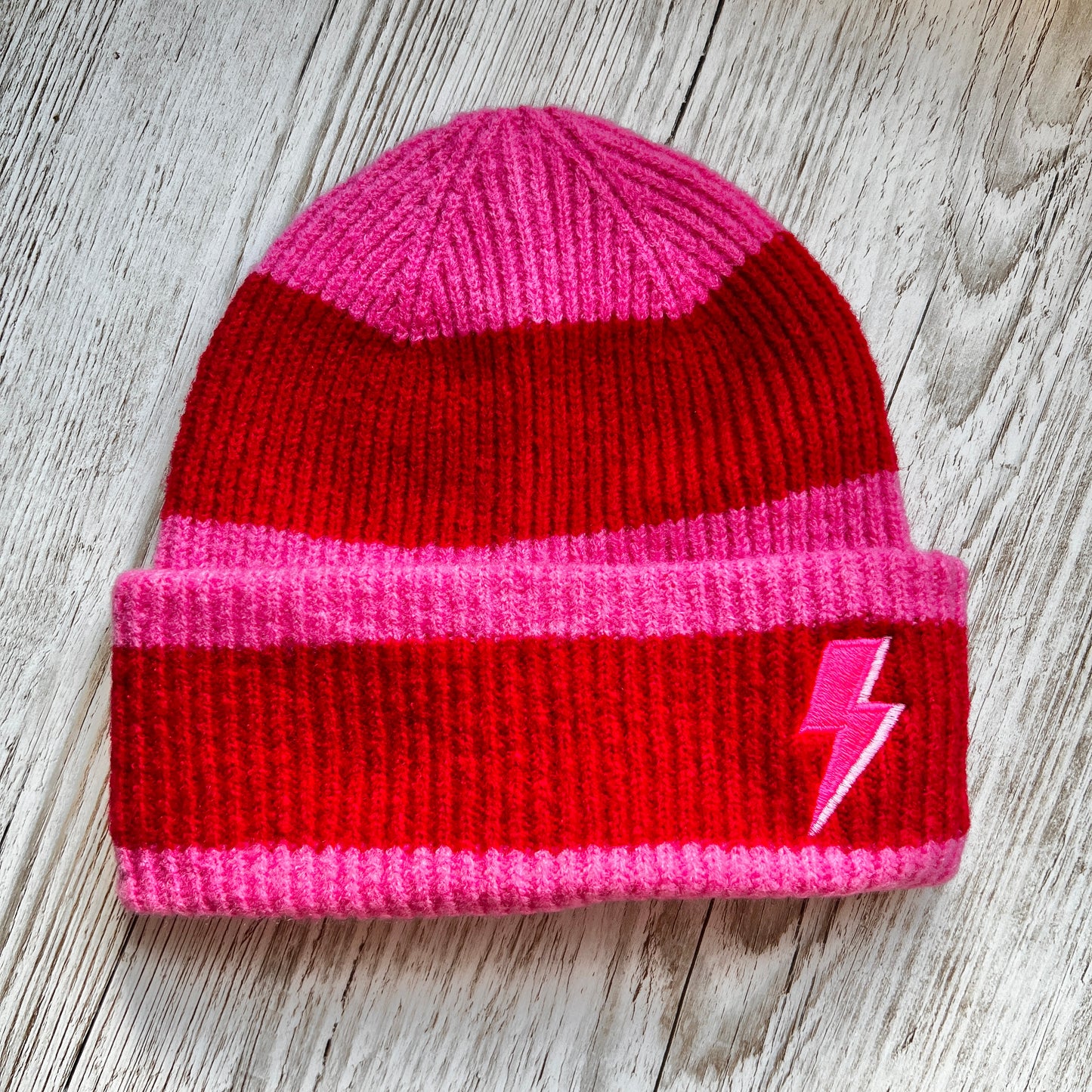 Red and pink ribbed beanie with a pink Embroidered lightning bolt on the cuff, on a wooden back ground 