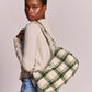 A model wearing a beige long-sleeve top and blue jeans is shown from the back and side, carrying a green and beige plaid shoulder bag against a solid light gray background.
