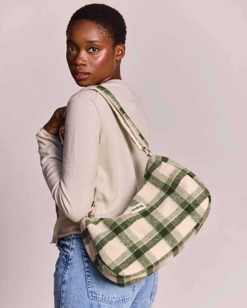 A model wearing a beige long-sleeve top and blue jeans is shown from the back and side, carrying a green and beige plaid shoulder bag against a solid light gray background.