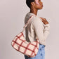 A woman wearing a beige long-sleeve top and blue jeans carries a slouchy shoulder bag made of a soft, textured fabric in a dusty pink and red plaid pattern, photographed against a solid light gray background.