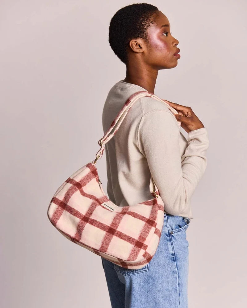 A woman wearing a beige long-sleeve top and blue jeans carries a slouchy shoulder bag made of a soft, textured fabric in a dusty pink and red plaid pattern, photographed against a solid light gray background.