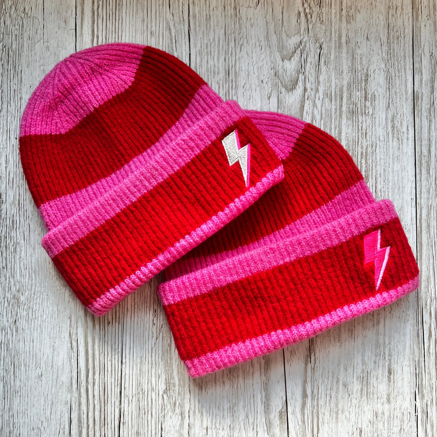 Two Red and pink stripped beanies with pink and silver embroidered bolts on a wooden background 