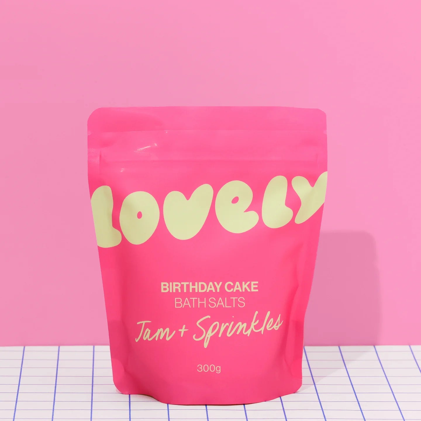 A pink stand-up pouch of Lovely Birthday Cake Bath Salts (Jam + Sprinkles, 300g) set against a solid pink background and a white grid-patterned surface.