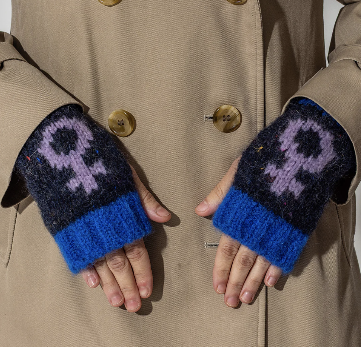Person wearing blue and black knitted fingerless gloves with a lilac female symbol design, against a beige coat background.