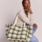 A woman kneels on a light gray studio floor wearing a beige long-sleeve top, blue jeans, and sneakers, while carrying a large cream canvas tote bag with a green plaid pattern. A small white brand label is visible on the front of the bag.