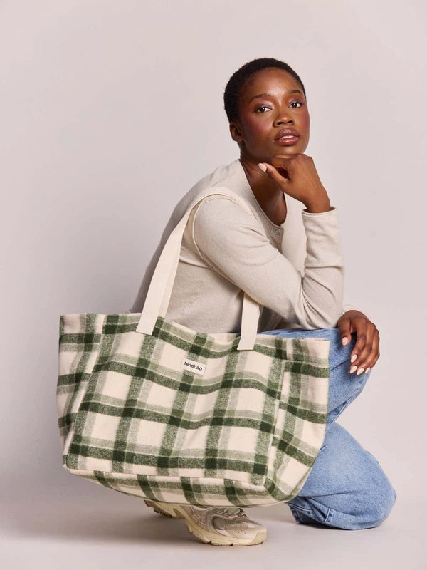 A woman kneels on a light gray studio floor wearing a beige long-sleeve top, blue jeans, and sneakers, while carrying a large cream canvas tote bag with a green plaid pattern. A small white brand label is visible on the front of the bag.