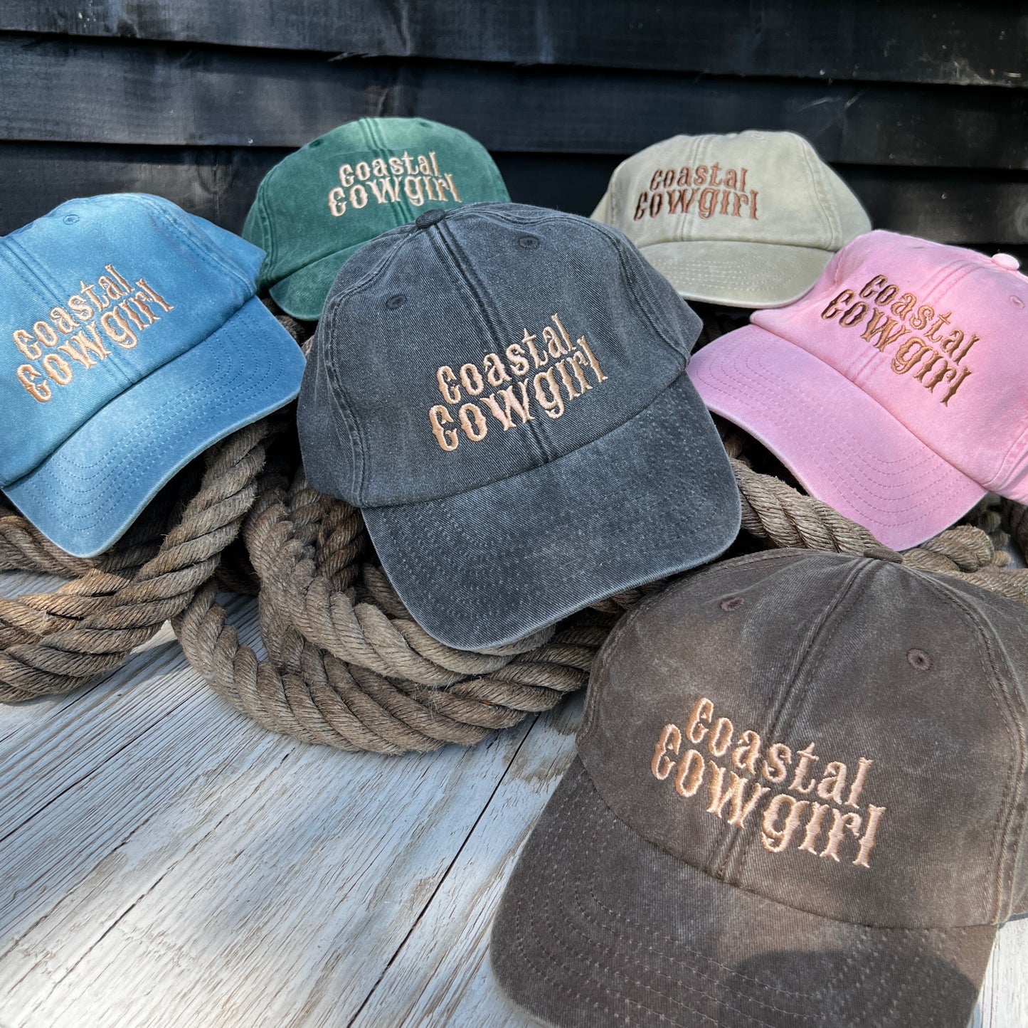 A collection of 'Coastal Cowgirl' branded caps in various colors, displayed in a stack with a rope, featuring the text embroidered on the front.