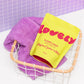 Yellow packet of 'lovely' elderflower bath salts with a purple towel in a basket against a purple tiled wall.