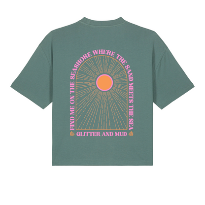 A green women's T-shirt with a graphic arch design featuring the text 'Find me on the seashore where the sand meets the sea, glitter and mud' and a pink sun motif.
