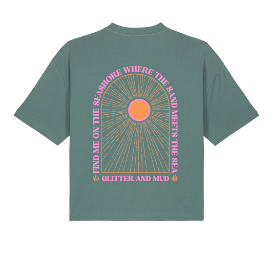 A green women's T-shirt with a graphic arch design featuring the text 'Find me on the seashore where the sand meets the sea, glitter and mud' and a pink sun motif.