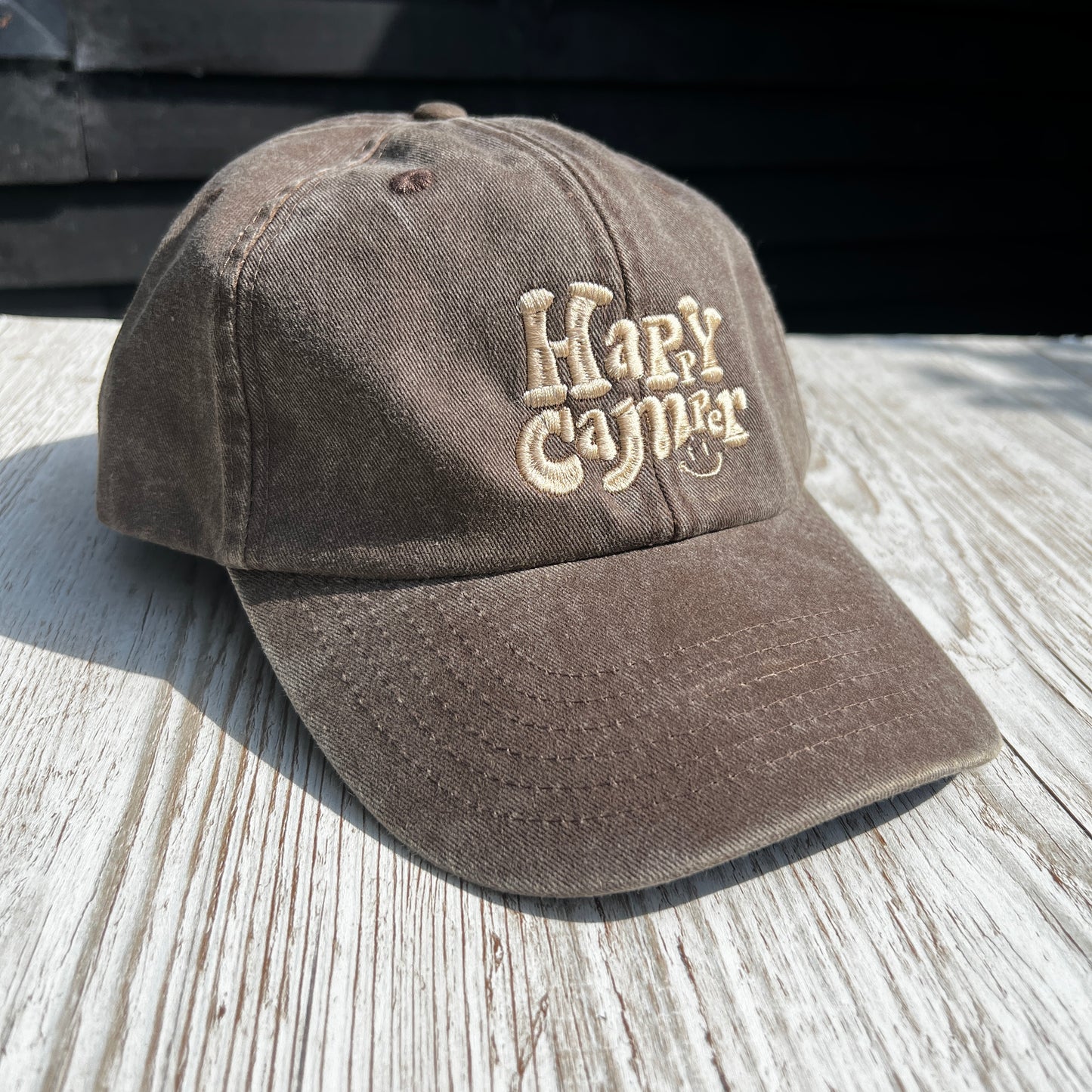 washed denim brown cap with beige embroidered 'Happy camper' on a light wooden surface 