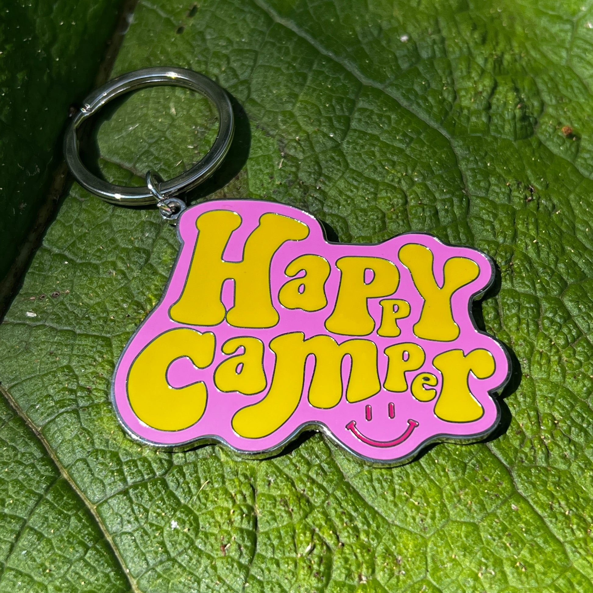 pink and yellow enamel 'Happy Camper' Key ring with a small red smiley face, on a green leaf 