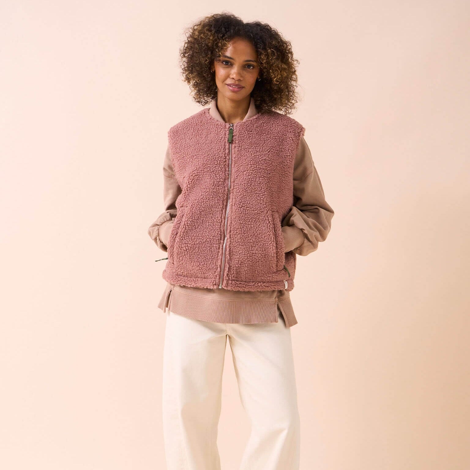 Woman wearing a pink fleece vest over a beige shirt with white pants on a beige background
