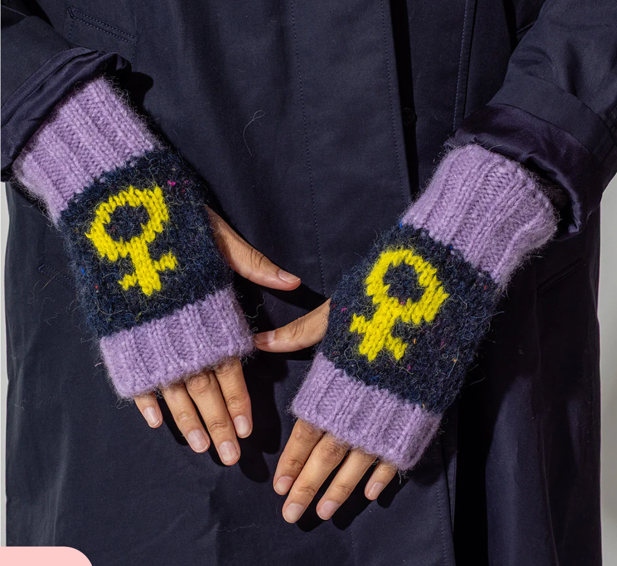 Purple and navy knitted fingerless gloves with yellow feminist symbols worn by a person in a dark coat