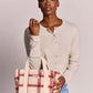 A woman wearing a beige long-sleeve button-up cardigan and light blue jeans holds a cream-colored plaid tote bag with red stripes. The bag features a small white label with the brand name 'handily'.