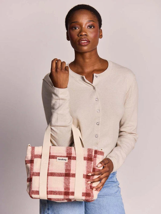 A woman wearing a beige long-sleeve button-up cardigan and light blue jeans holds a cream-colored plaid tote bag with red stripes. The bag features a small white label with the brand name 'handily'.
