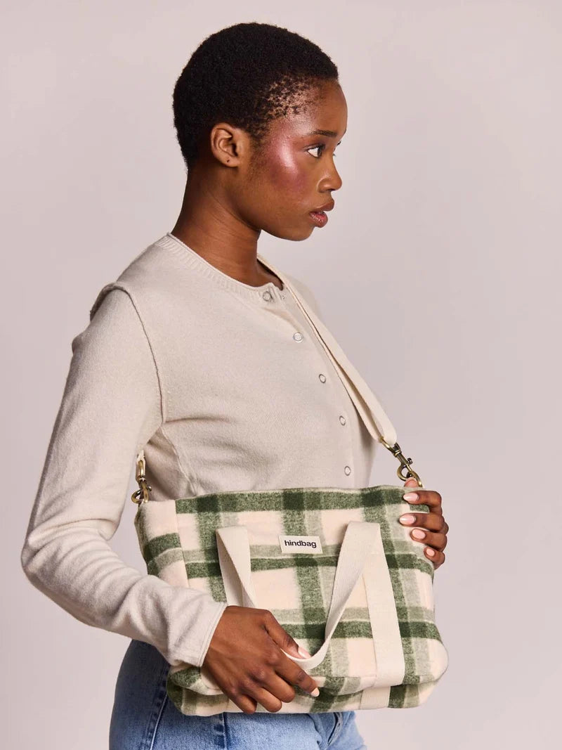 A woman wearing a cream cardigan and blue jeans holds a green and cream plaid tote bag with a shoulder strap. The bag has a small white label that reads 'hindbag'.