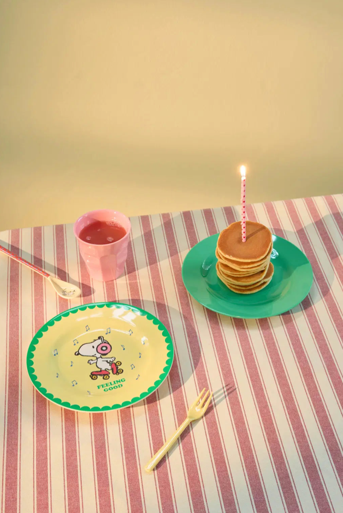 A styled tabletop scene with a stack of pancakes on a green plate with a single lit candle, a printed round plate with a cartoon character, a pink cup, and a small fork on a pink-and-white striped tablecloth.