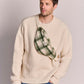 A man wearing a cream ribbed knit crewneck sweater and blue jeans, carrying a green and beige plaid fanny pack/bum bag across his chest.