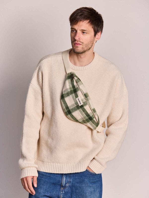 A man wearing a cream ribbed knit crewneck sweater and blue jeans, carrying a green and beige plaid fanny pack/bum bag across his chest.