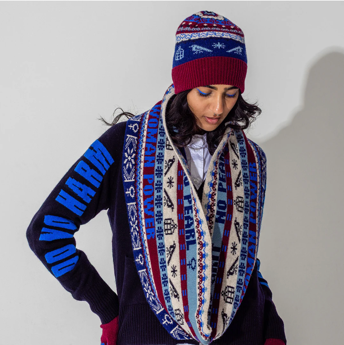 Person wearing a patterned beanie and scarf against a plain background blue, red and beige knitted snood 'No Grit No Pearl, Woman Power' text and symbols on a white background