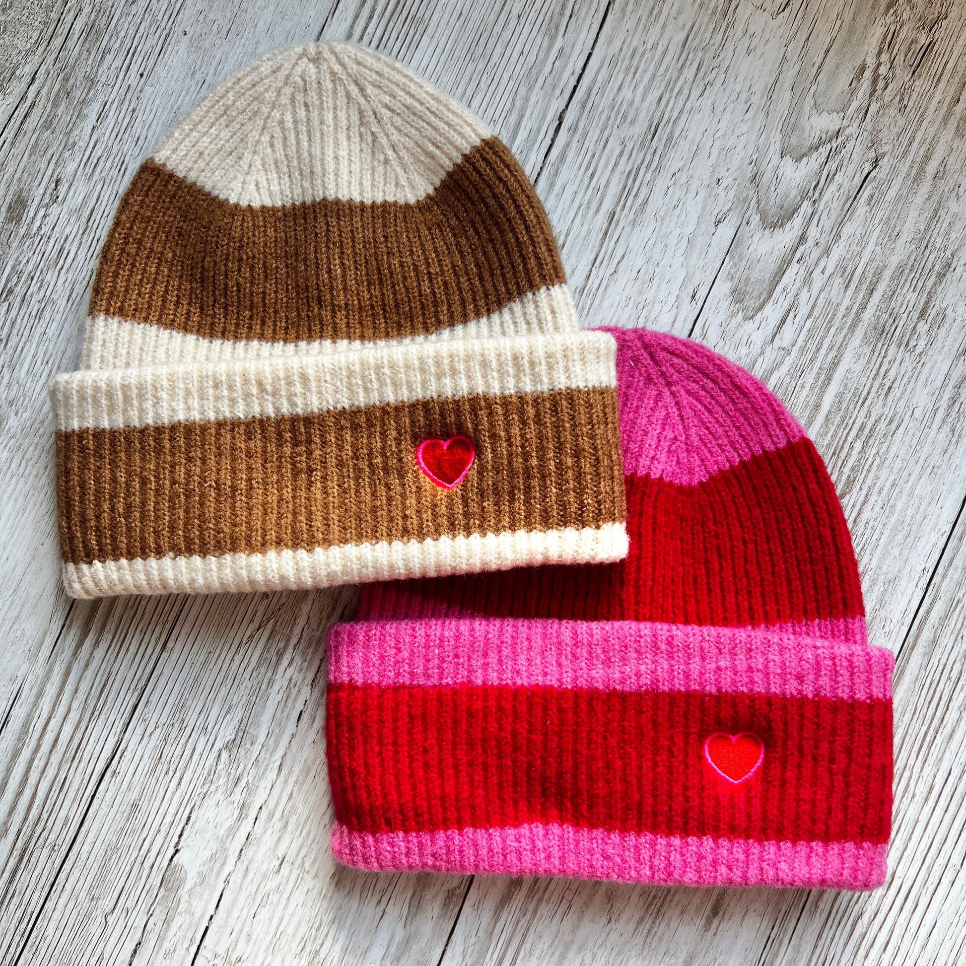 two stripped beanie hats laying on a wooden background. a brown and beige striped hat is on top of a pink and red striped hat both embroidered with a red heart on the cuff. 